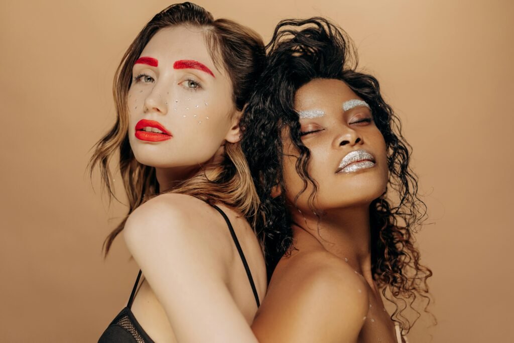 Glamorous studio portrait of two women with vibrant glitter makeup and stylish poses.